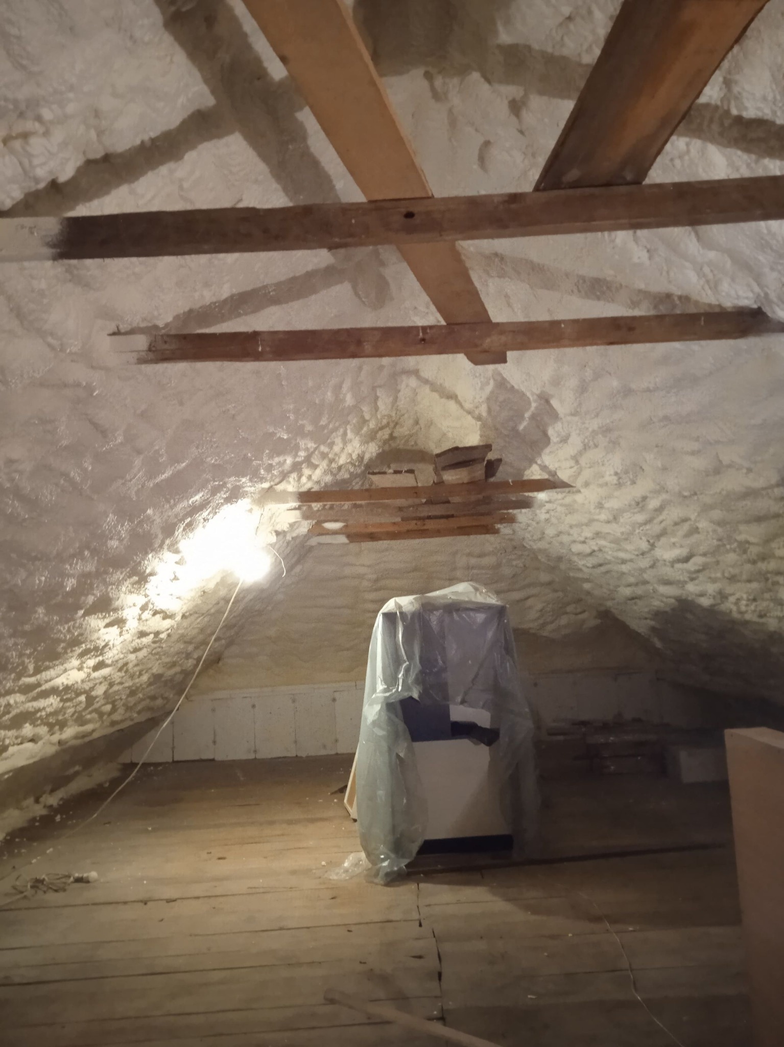 Poddasze z widoczną warstwą izolacji natryskowej z pianki PUR, drewnianymi belkami stropowymi i zakrytym urządzeniem, prawdopodobnie agregatem, folią ochronną.