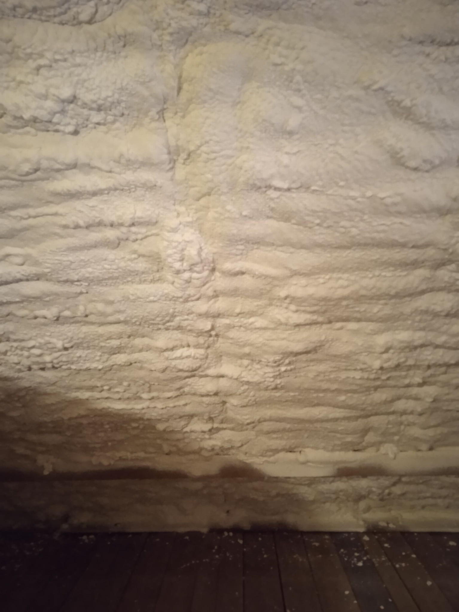 Poddasze po natrysku pianki poliuretanowej, widoczna nierówna struktura izolacji i fragment drewnianej podłogi w dolnej części kadru.