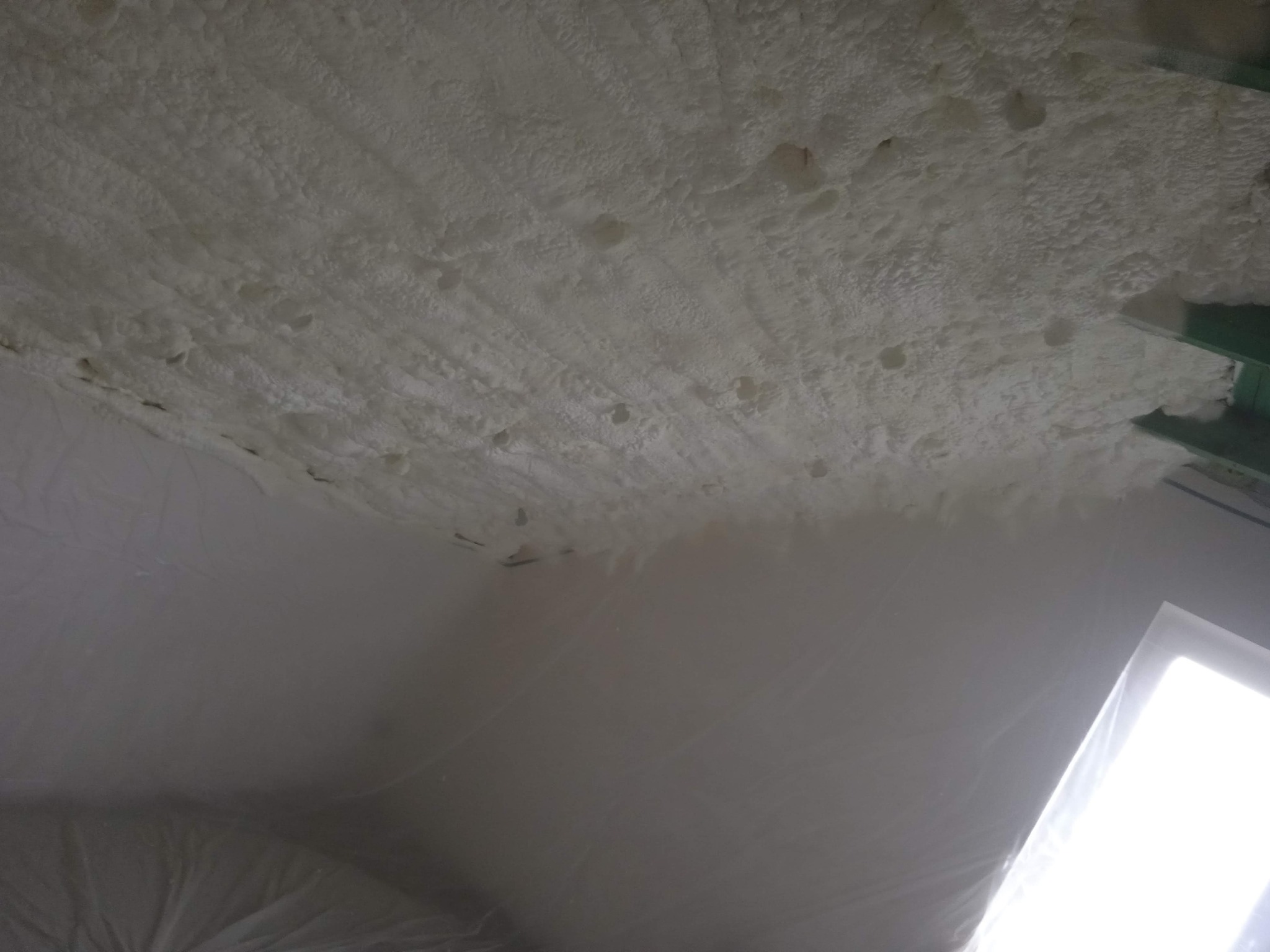 Nierównomierna warstwa białej pianki izolacyjnej naniesiona na sufit poddasza, widoczne fragmenty konstrukcji dachu i folii ochronnej.