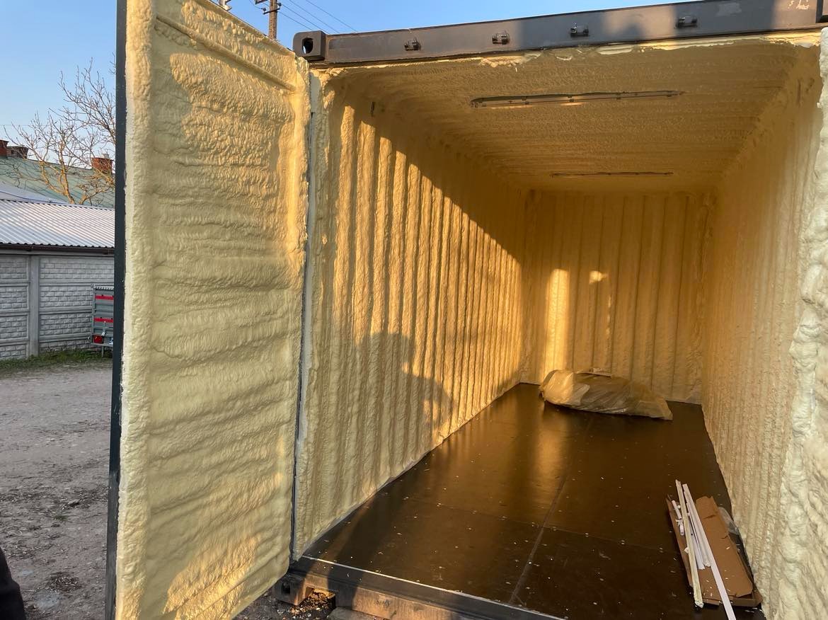 Wnętrze kontenera po natrysku pianką poliuretanową, widoczne równomierne pokrycie ścian, sufitu i otwartych drzwi, podłoga wyłożona panelami, w tle budynki mieszkalne.