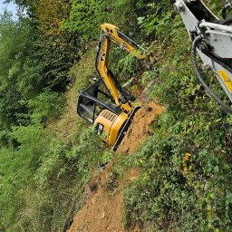 M-KOP Prace Ziemne Mateusz Sobczyk - Żółta mini koparka CAT na zboczu porośniętym zielenią, w tle fragment innej maszyny i siedzący pracownik w kamizelce odblaskowej.
