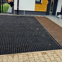 M-KOP Prace Ziemne Mateusz Sobczyk - Pracownik z łopatą ubija ziemię w kratce trawnikowej na podjeździe, obok ułożona kostka brukowa i fragment domu z oknem oraz worki z materiałem.