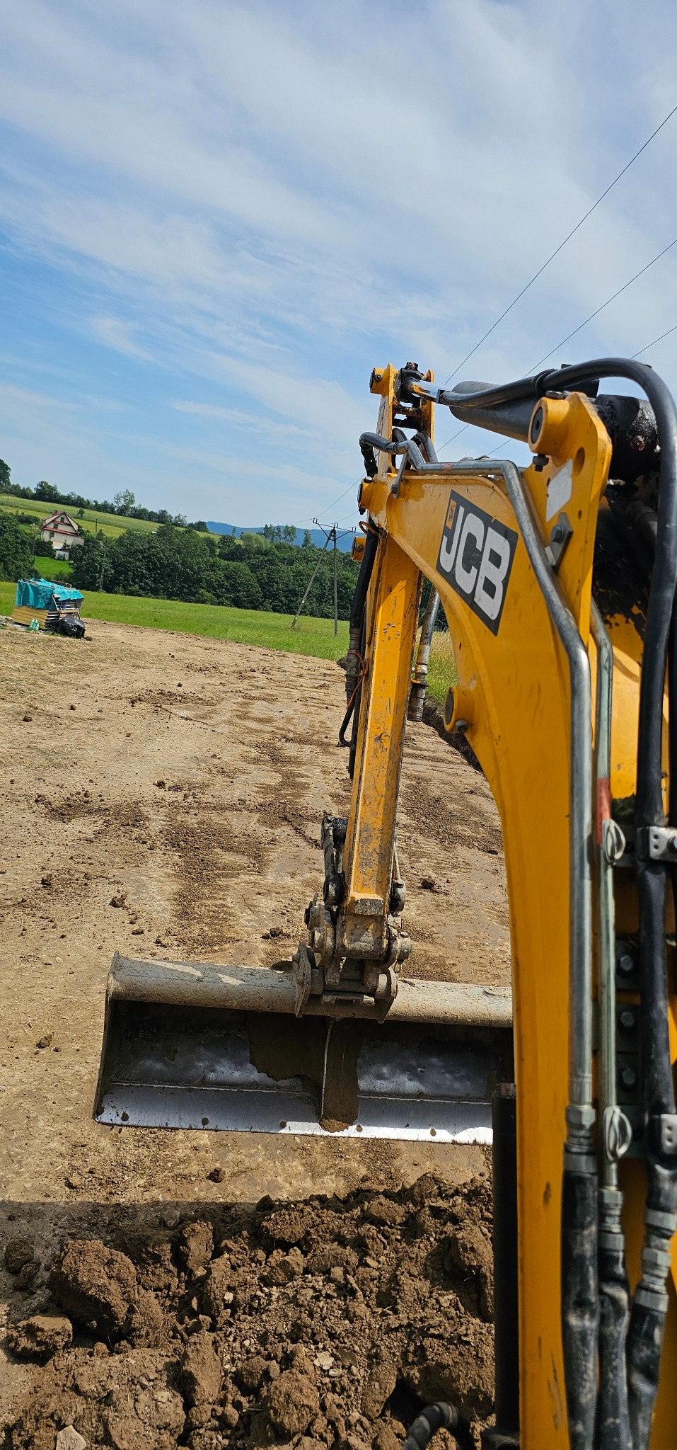 Żółta koparka JCB w trakcie pracy na polu, widoczne świeżo przekopane grudki ziemi, w tle zielone wzgórza i błękitne niebo z chmurami.
