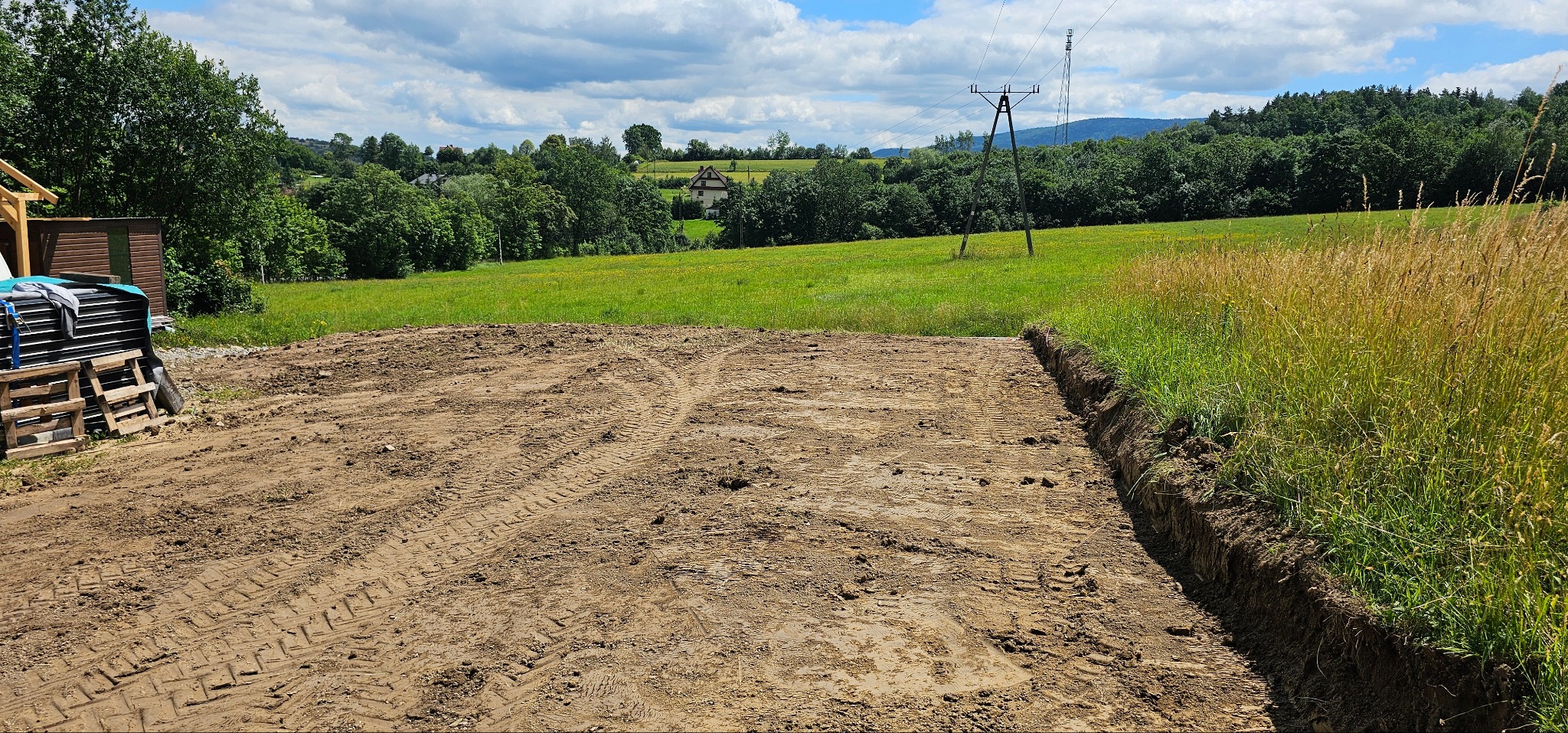 Przygotowany teren pod budowę z widocznymi śladami opon, wykopem wzdłuż krawędzi i stertą materiałów budowlanych na paletach w tle, malowniczy krajobraz z polami i lasami w oddali.