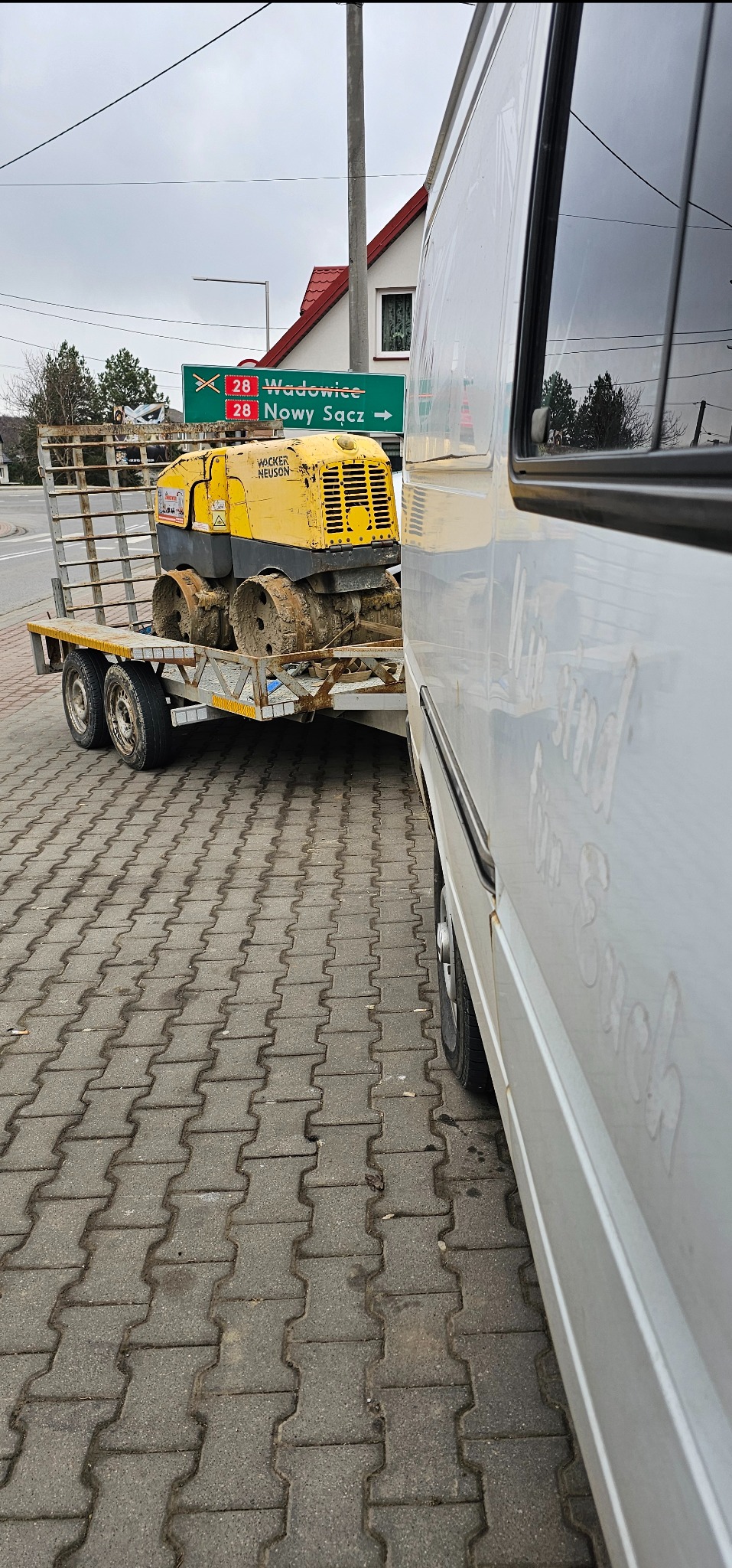 Żółty walec wibracyjny Wacker Neuson na przyczepie lawetowej, częściowo zasłonięty przez biały samochód dostawczy, widoczny znak drogowy z nazwami miejscowości: Wadowice i Nowy Sącz.