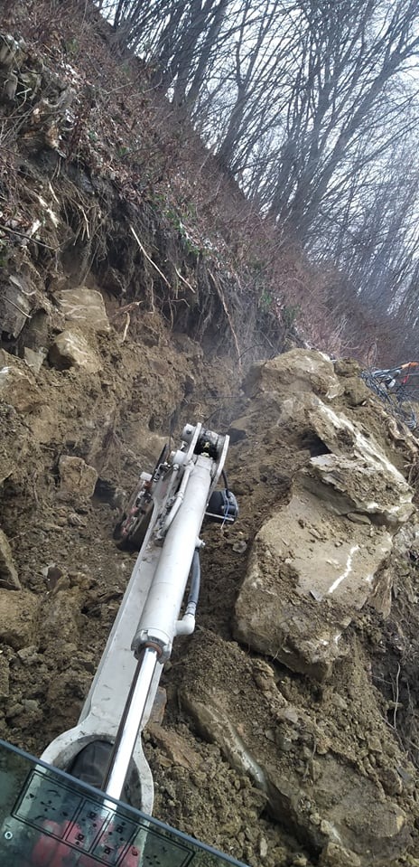 Ramię koparki hydraulicznej wgryza się w ziemię na zboczu porośniętym drzewami, przygotowując wykop pod instalacje.