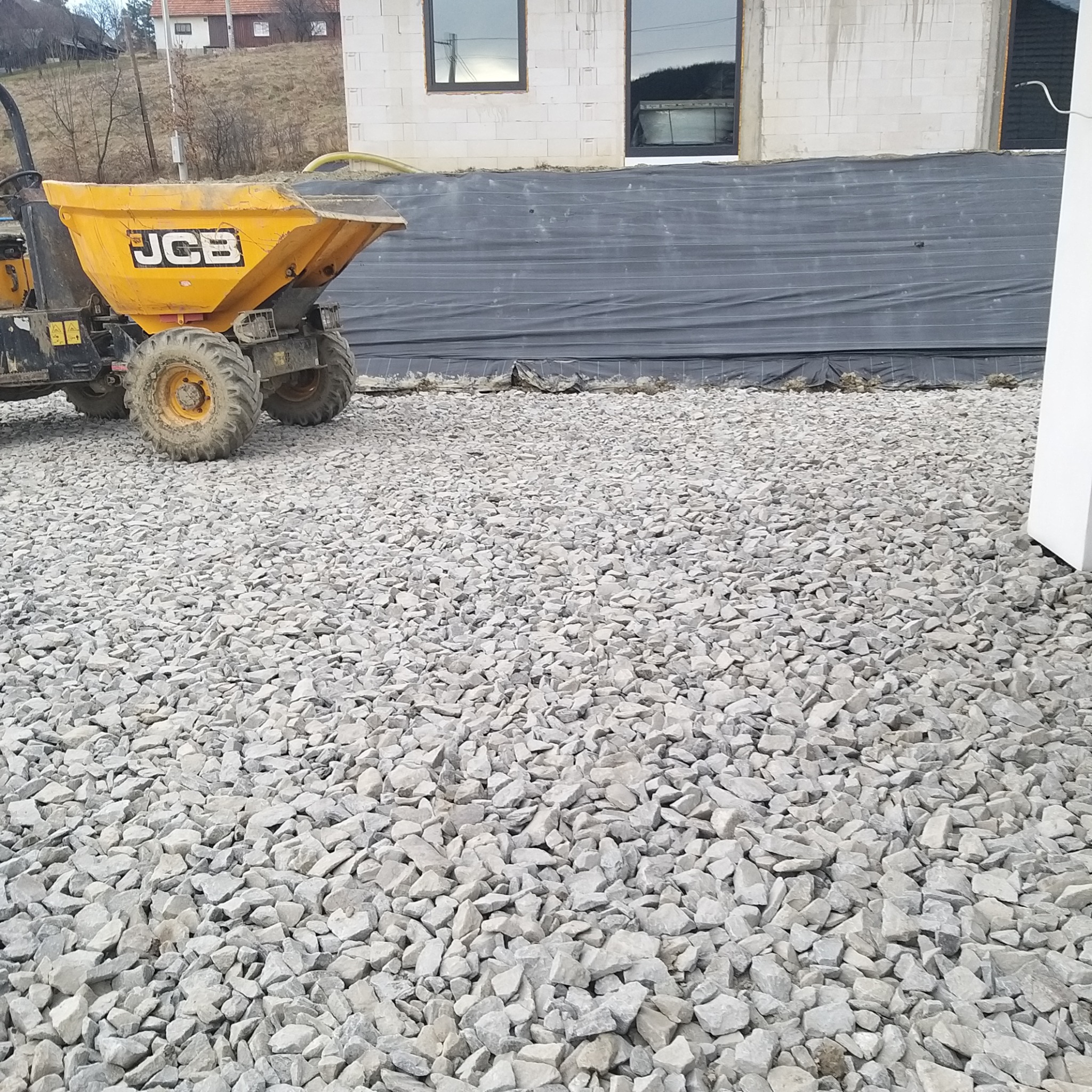 Żółta wywrotka JCB na tle nowo wznoszonego budynku z oknami, świeżo utwardzona droga z szarego kruszywa.