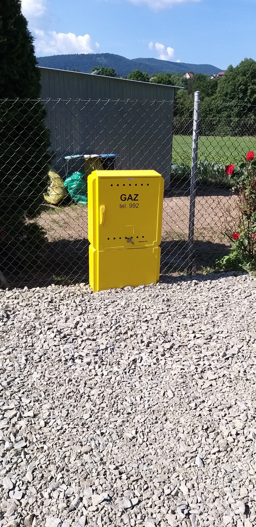 Żółta szafka gazowa z napisem 'GAZ tel. 992' umieszczona na żwirowym podłożu, widoczna przez siatkę ogrodzeniową, w tle budynek gospodarczy, drzewa i wzgórza w słoneczny dzień.