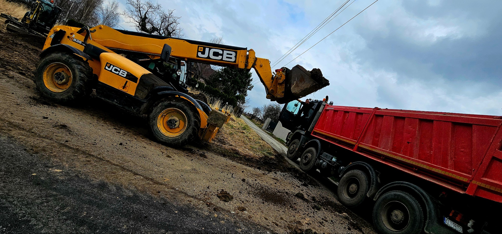 Żółta ładowarka JCB nabiera ziemię i wysypuje ją do czerwonej ciężarówki na placu budowy, pochmurne niebo w tle.