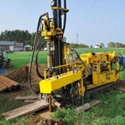 Żółta wiertnica Beretta T26 w trakcie pracy na zielonym polu, widoczne rury wiertnicze i budynek gospodarczy w tle.