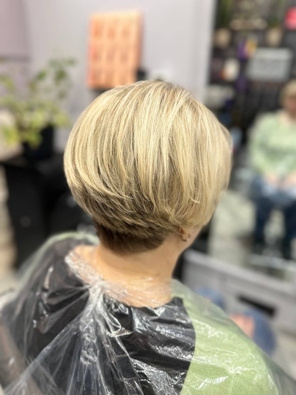 Blond włosy ścięte w boba, widok z tyłu, klientka w pelerynie fryzjerskiej, widoczne delikatne cieniowanie przy karku.