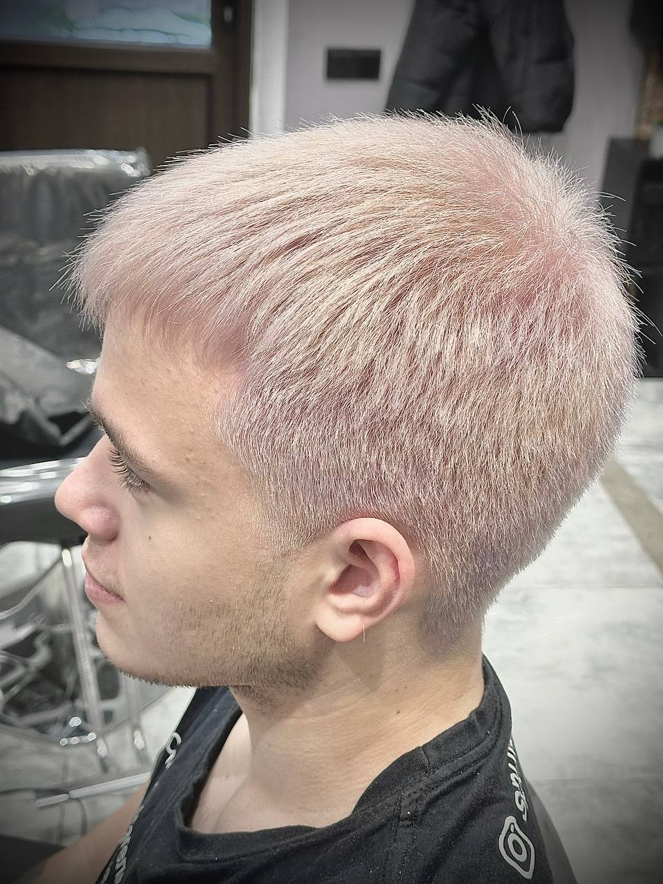 Młody mężczyzna z krótkimi, platynowo-różowymi włosami, widok z profilu, ukazujący świeżo ostrzyżoną fryzurę i delikatny kolczyk w uchu.