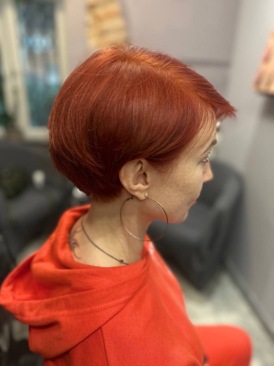 Krótkie, równo ostrzyżone włosy w odcieniach miedzi, widoczne z profilu. Modelka ma na sobie czerwoną bluzę z kapturem i duże, okrągłe kolczyki.