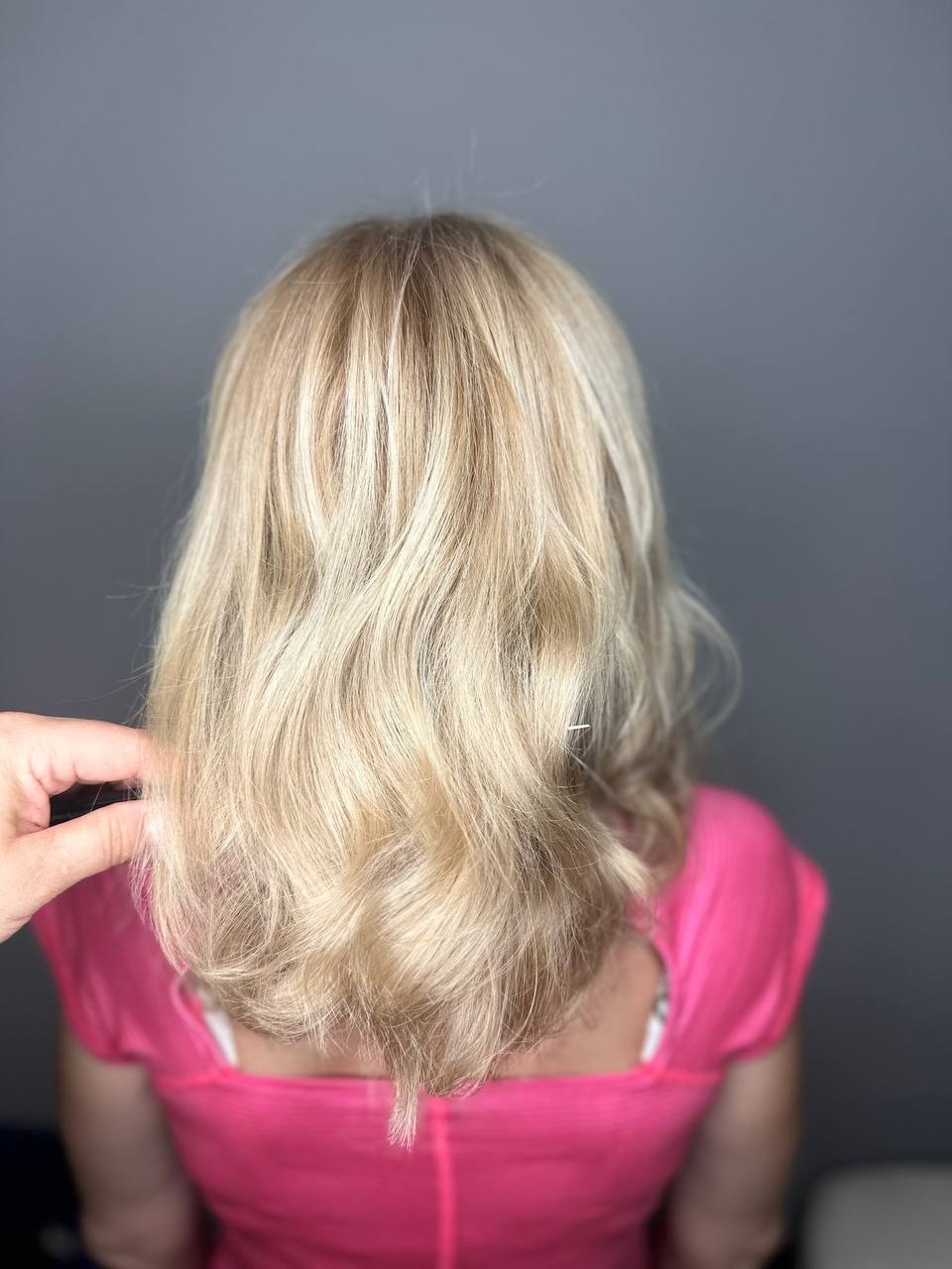 Blond włosy po koloryzacji z delikatnymi falami, widok z tyłu, z ręką dotykającą pasma po lewej stronie.