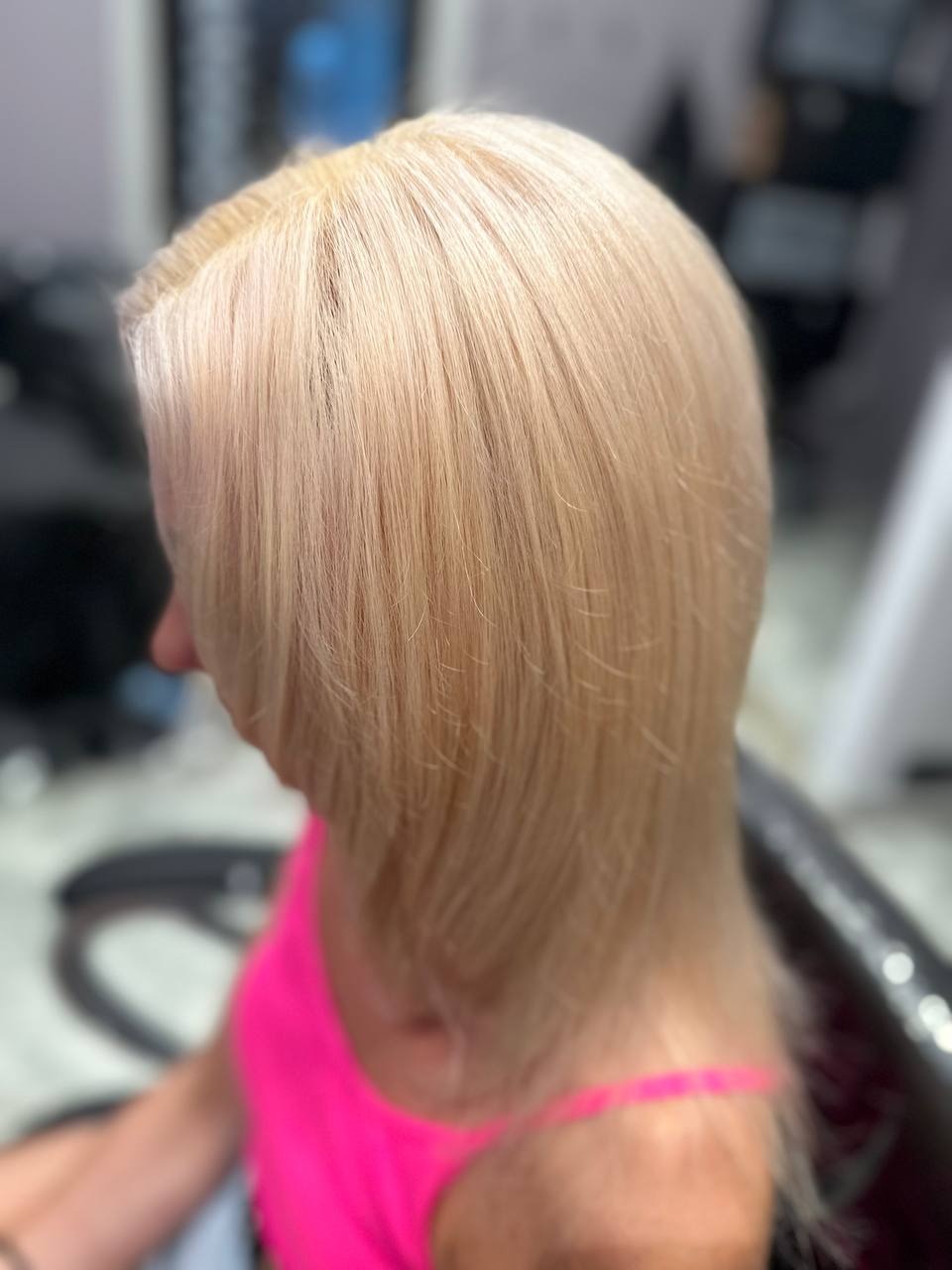 Blond włosy kobiety po koloryzacji, widoczne od tyłu z profilu, w różowej koszulce, na fotelu w salonie.