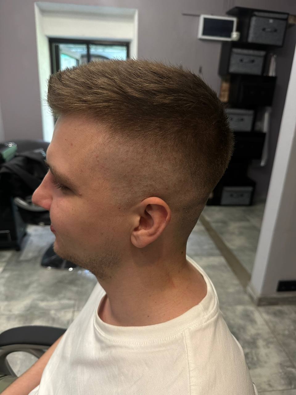 Męska fryzura z krótkimi bokami i dłuższym wierzchem, widok z profilu, z delikatnym cieniowaniem, na tle wnętrza salonu.