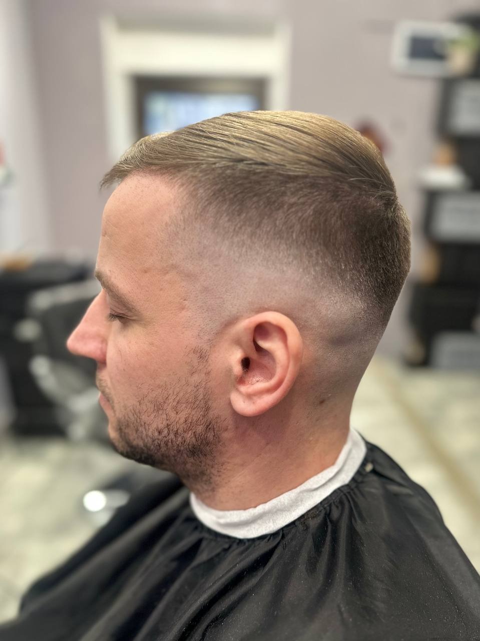 Mężczyzna z krótką, precyzyjnie wycieniowaną fryzurą typu fade, widoczny z profilu, w czarnej pelerynie ochronnej.