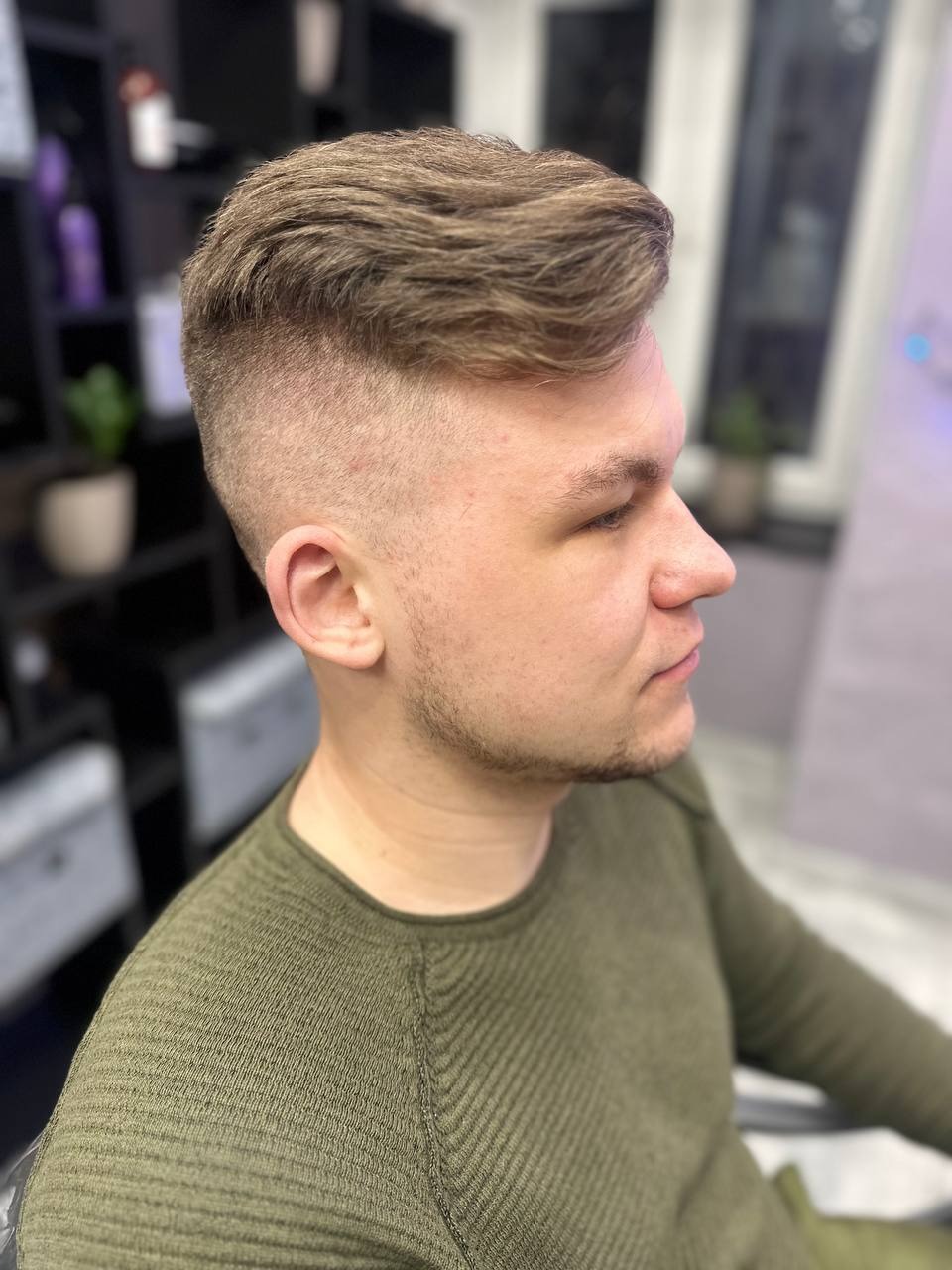 Męska fryzura z cieniowaniem na bokach i dłuższa góra, widok z profilu, klient w zielonym swetrze.