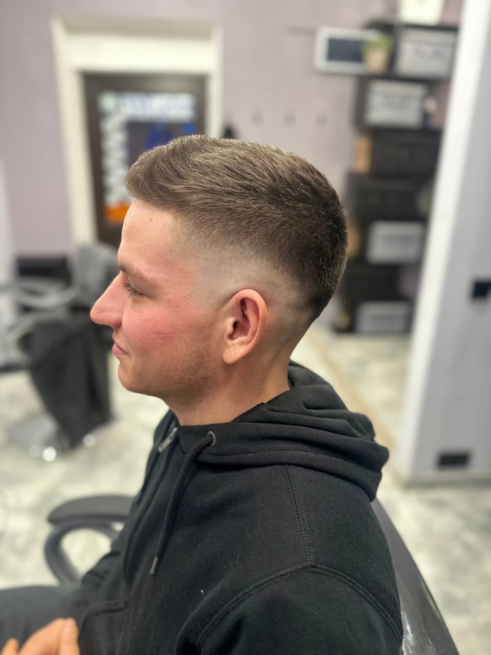 Męska fryzura z krótkim cieniowaniem na bokach i dłuższymi włosami na górze głowy, widok z profilu, klient ubrany w czarną bluzę z kapturem siedzi na fotelu.