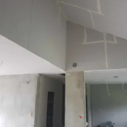 Wnętrze w trakcie montażu płyt kartonowo-gipsowych na skośnym suficie, widoczne szpachlowane łączenia, otwory na oświetlenie i wystające kable.