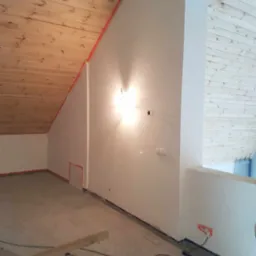 Poddasze w trakcie remontu: drewniana boazeria na skośnym suficie, pomalowane ściany z zabezpieczeniami taśmą malarską, widoczne gniazdka elektryczne i przewody na podłodze.