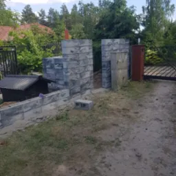 Wznoszenie ogrodzenia z szarych, betonowych bloczków imitujących kamień, widoczna brama wjazdowa i fragment zabudowy mieszkalnej w tle.
