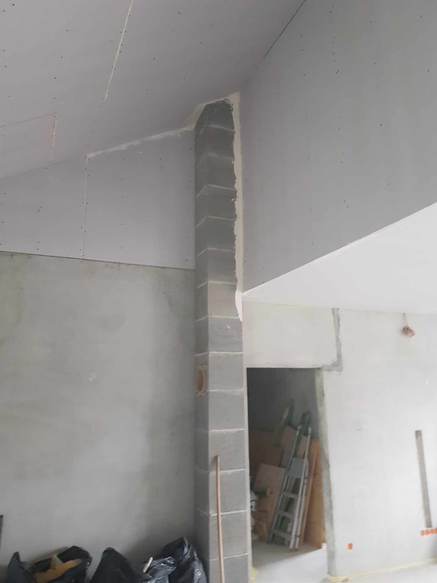 Wnętrze w trakcie prac wykończeniowych: sufit i ściany częściowo pokryte płytami kartonowo-gipsowymi, fragment kolumny z surowych bloczków, widoczne worki z odpadami i drabina w przejściu do innego...