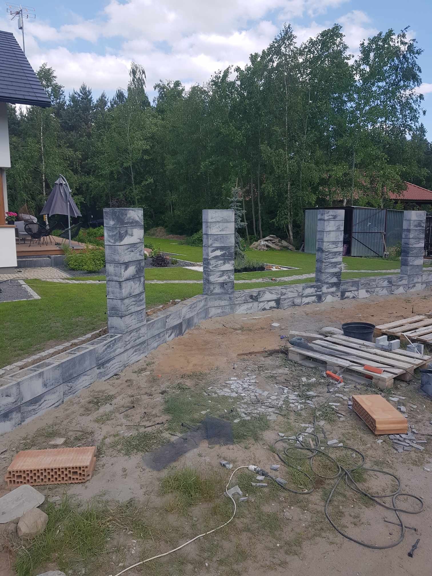 Wznoszenie ogrodzenia z betonowych bloczków o marmurkowym wzorze, widoczne palety z narzędziami i materiałami budowlanymi na niezagospodarowanym terenie, w tle dom z tarasem i zielony ogród.