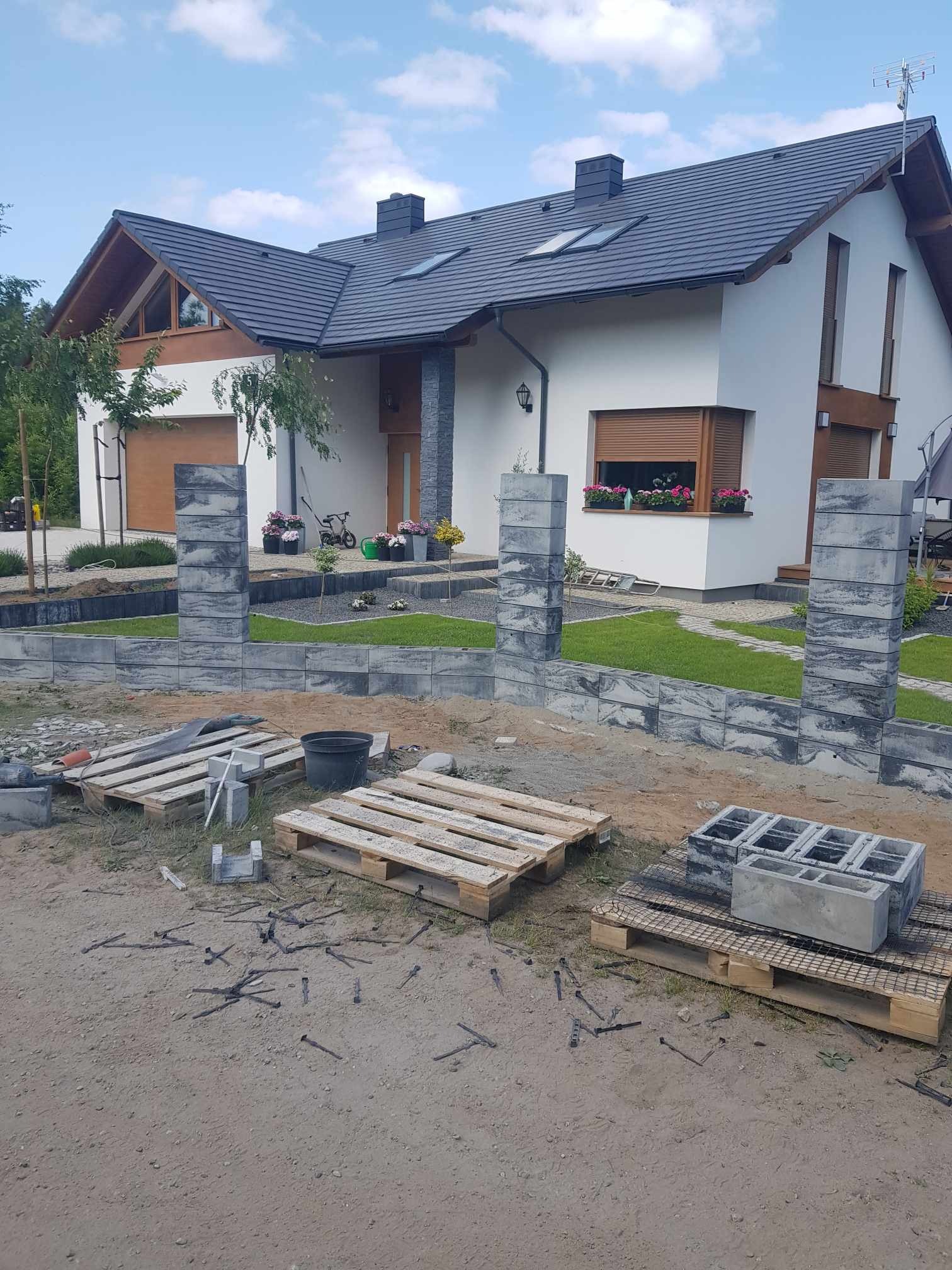 Budowa niskiego murku z szarych bloczków betonowych przed domem jednorodzinnym z białymi ścianami i szarą dachówką, widoczne palety z materiałami budowlanymi i rozsypane pręty zbrojeniowe na ziemi.