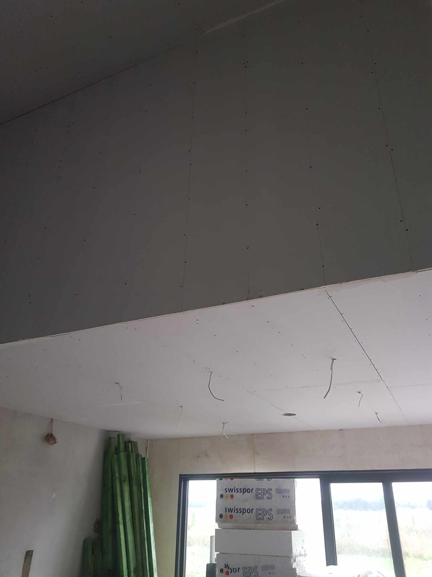 Wnętrze w trakcie prac wykończeniowych: widoczne płyty kartonowo-gipsowe na suficie, częściowo pomalowane na szaro, z wystającymi przewodami elektrycznymi, zielone drewniane listwy opierające się...