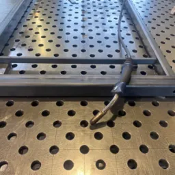 Metalowa rama spawana na stole spawalniczym z widocznym uchwytem spawalniczym i zaciskiem mocującym.