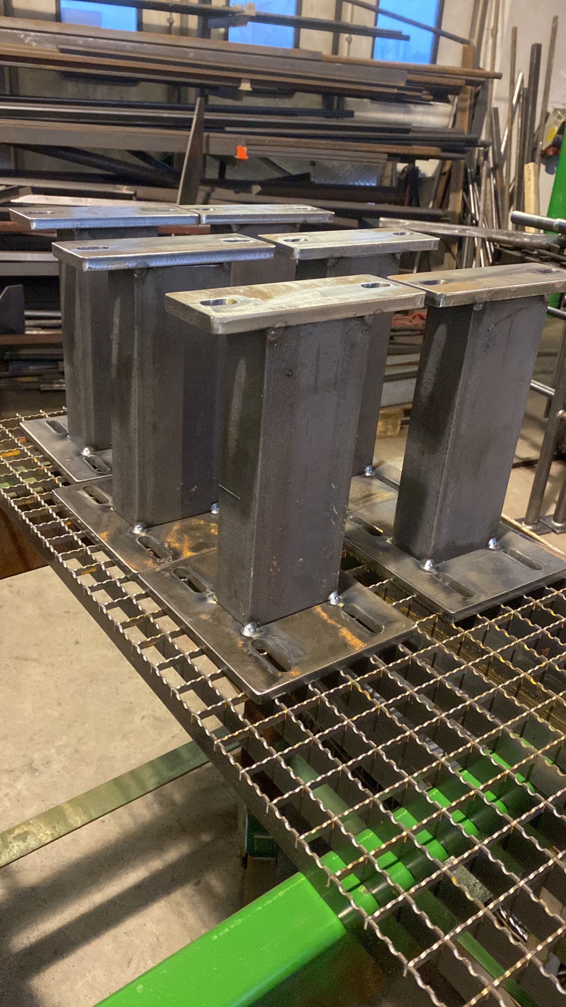 Surowe, metalowe słupki z przyspawanymi płytami, przygotowane do dalszej obróbki, umieszczone na metalowej kracie w warsztacie.