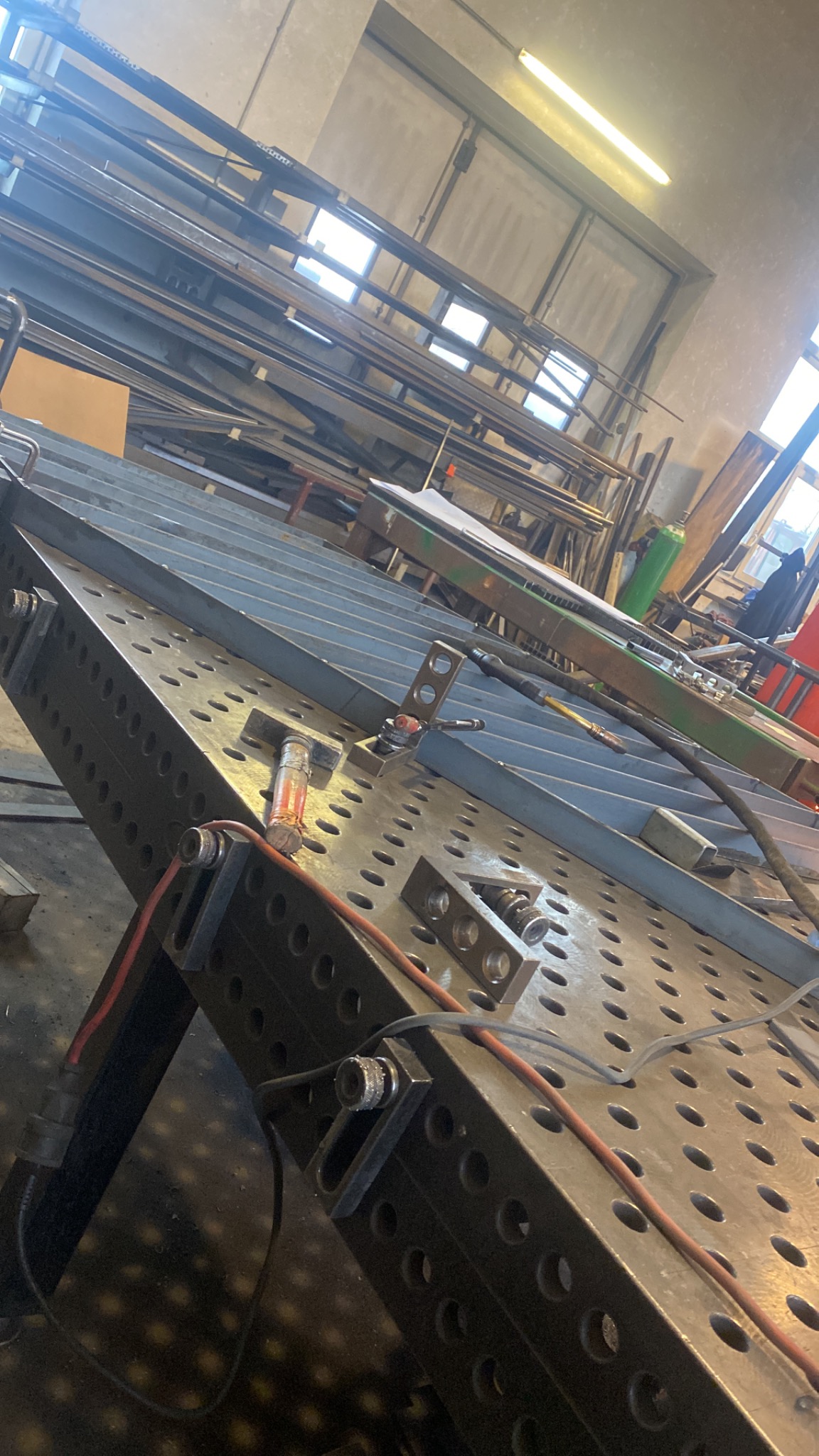 Stół spawalniczy z zamocowanymi elementami metalowymi, przygotowany do spawania, w tle widoczne profile i elementy konstrukcyjne w warsztacie.