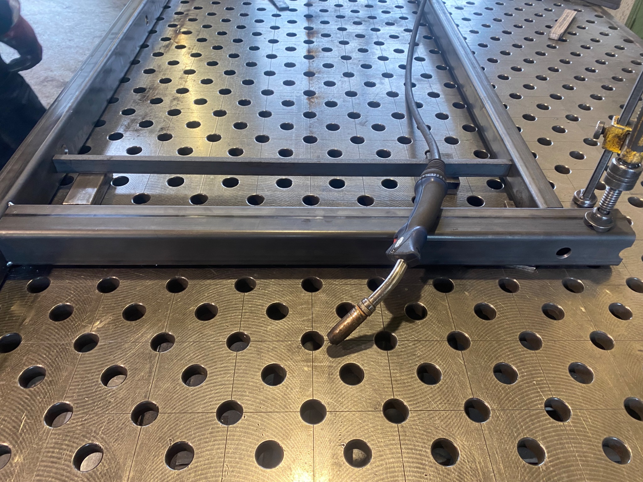 Metalowa rama spawana na stole spawalniczym z widocznym uchwytem spawalniczym i zaciskiem mocującym.