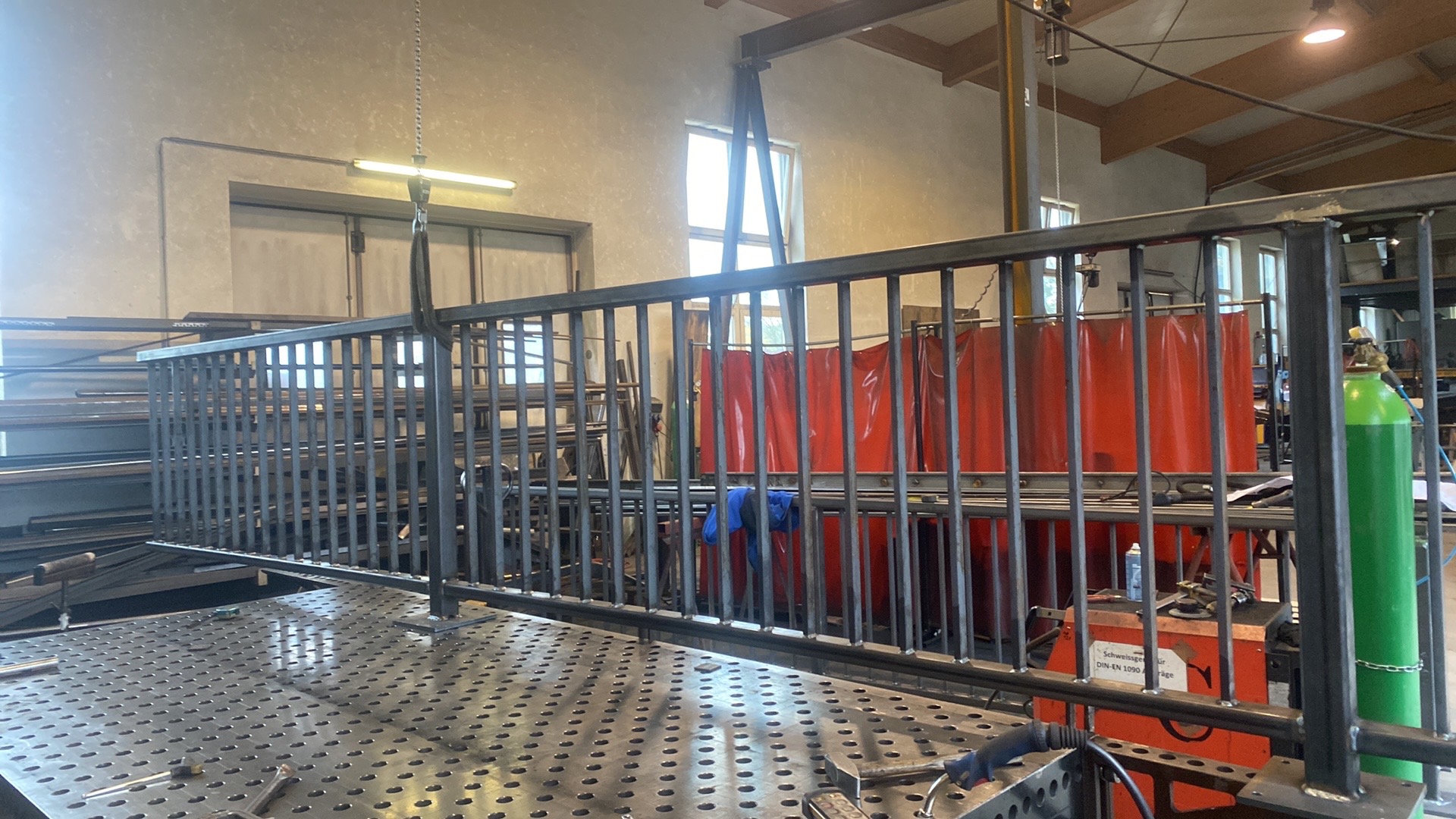 Metalowa konstrukcja balustrady w warsztacie, zawieszona na haku, z widocznym sprzętem spawalniczym i butlą z gazem w tle, na perforowanym stole warsztatowym.