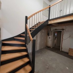 Patrycja Miszczak - Spiralne schody z jasnego drewna z czarnymi bokami i metalowymi balustradami prowadzące na antresolę; widoczny surowy stan wykończenia wnętrza.