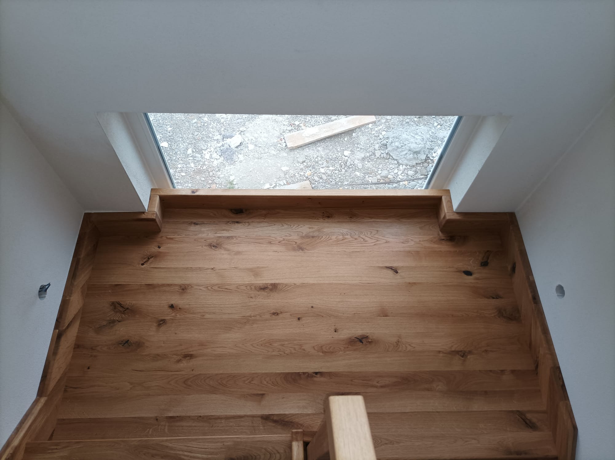 Widok z góry na drewniane schody z wyraźnym usłojeniem, w tle okno z widokiem na gruzowisko.