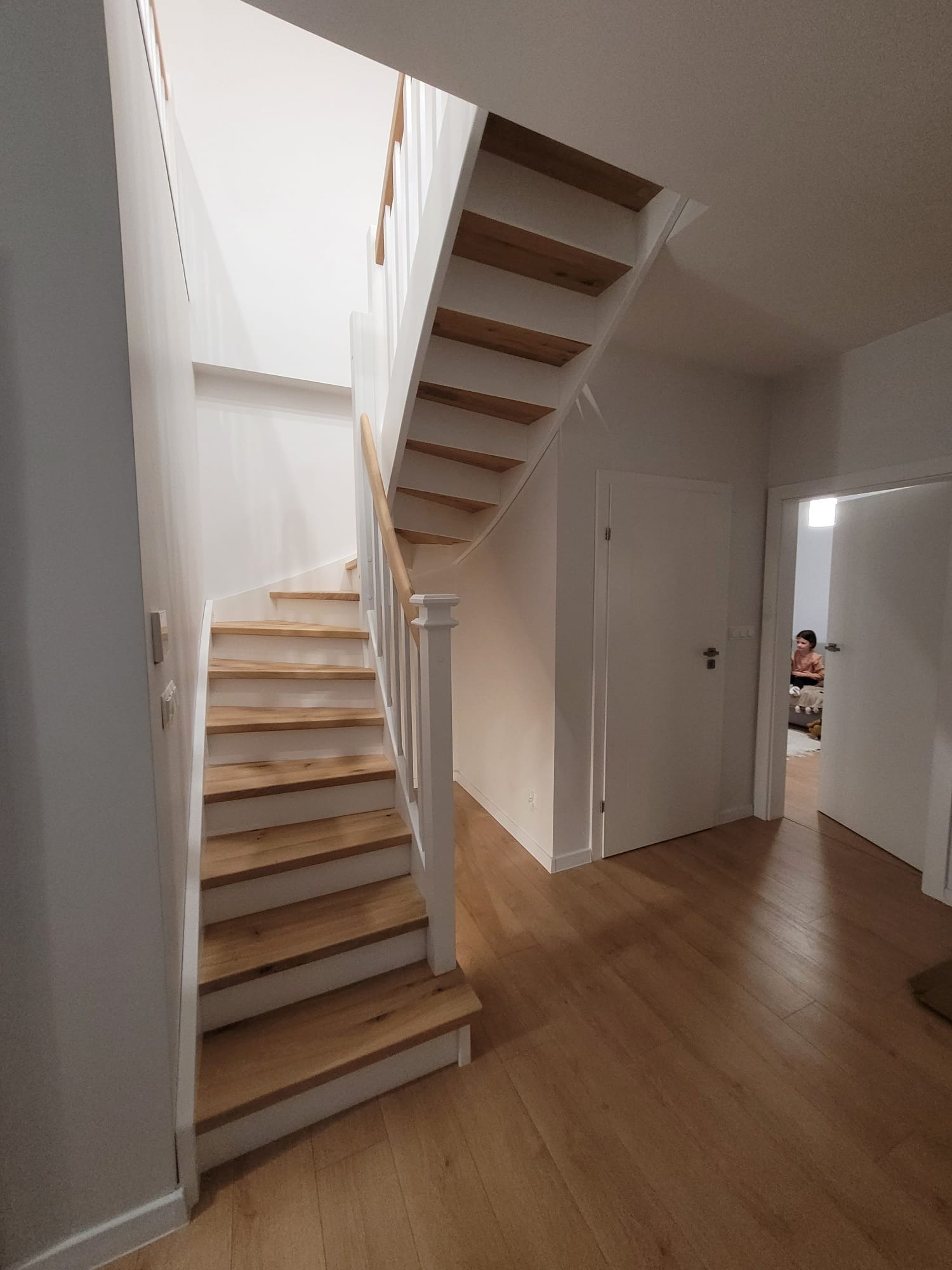 Jasne, drewniane stopnie schodów z białymi podstopnicami i balustradą, prowadzące do góry w nowoczesnym wnętrzu z drewnianą podłogą.