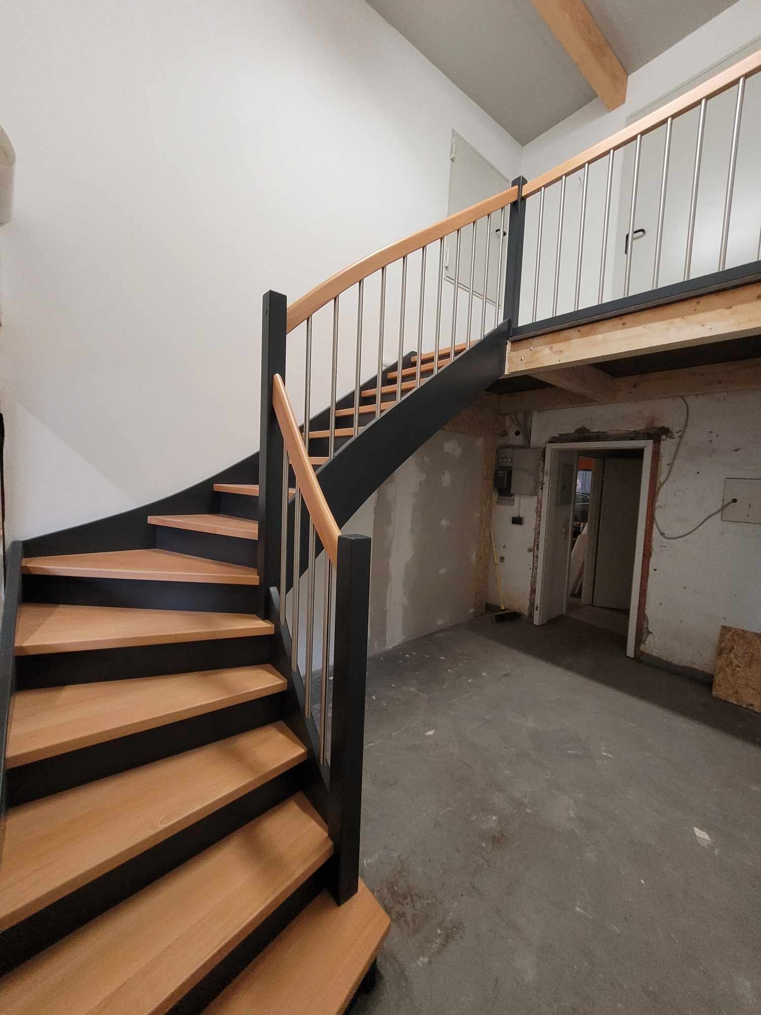 Spiralne schody z jasnego drewna z czarnymi bokami i metalowymi balustradami prowadzące na antresolę; widoczny surowy stan wykończenia wnętrza.