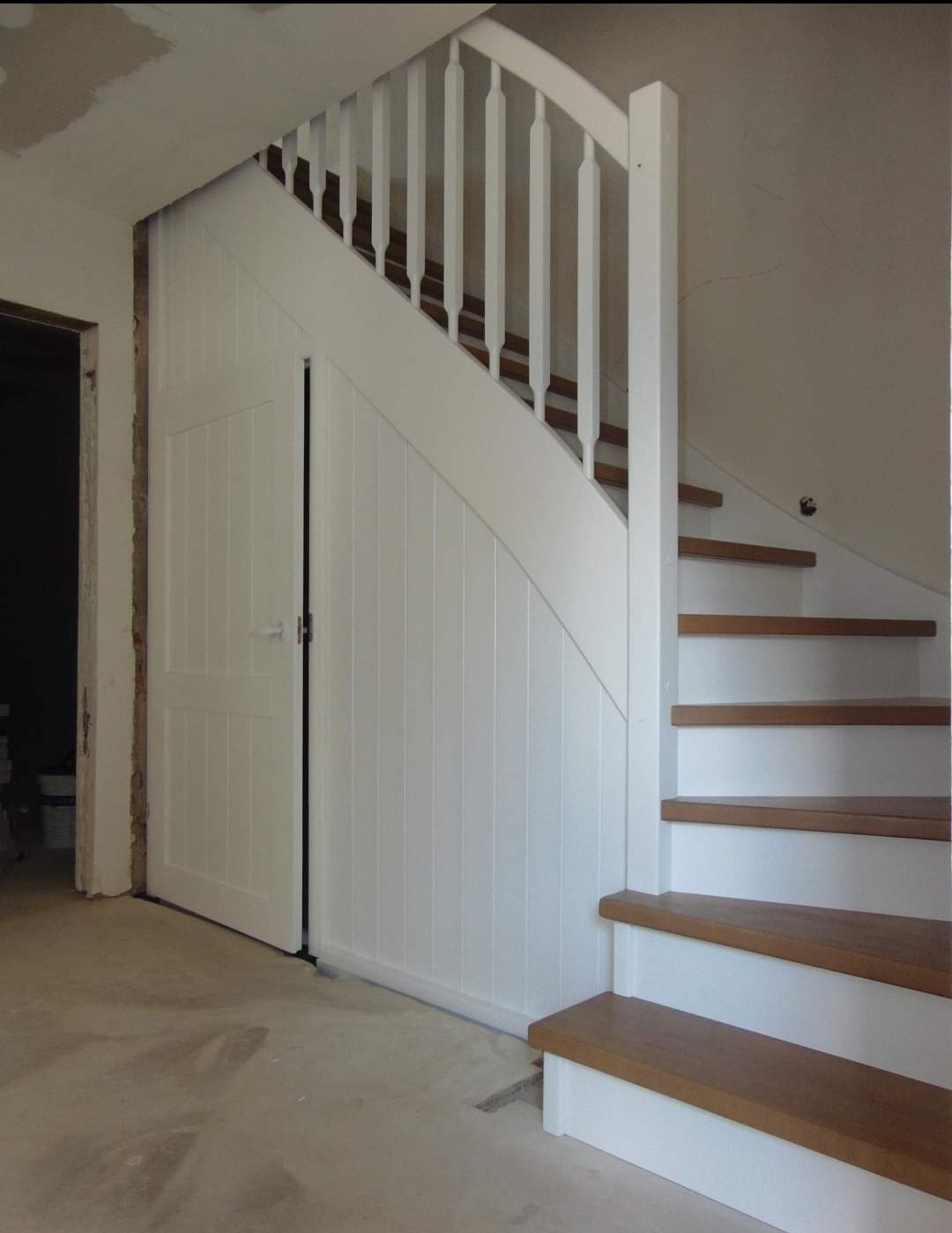 Białe schody z drewnianymi stopniami i balustradą, zabudowane z boku szafką z drzwiami panelowymi, widoczne niedokończone wykończenie ścian.