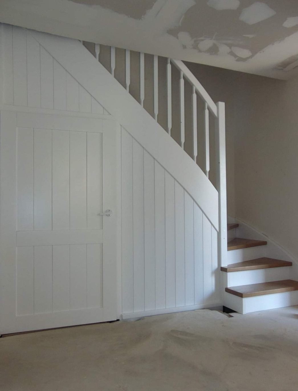 Białe schody z drewnianymi stopniami i białą balustradą, z zabudową pod schodami z drzwiami, surowy stan wykończenia pomieszczenia.