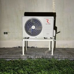 DKInstall - serwis urządzeń grzewczych i chłodniczych, montaż klimatyzacji - Biała jednostka zewnętrzna klimatyzatora Rotenso Unico Series, umieszczona na metalowym stelażu, stoi na brukowanym podłożu przed ścianą z widocznymi śladami wilgoci i instalacji.