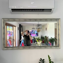DKInstall - serwis urządzeń grzewczych i chłodniczych, montaż klimatyzacji - Biały klimatyzator ścienny zamontowany nad lustrem w srebrnej ramie, w którym odbija się osoba robiąca zdjęcie. W tle widoczny salon z roślinami i telewizorem.
