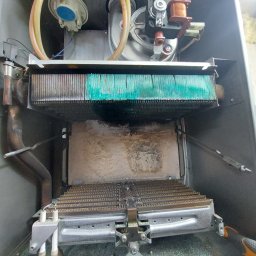 DKInstall - serwis urządzeń grzewczych i chłodniczych, montaż klimatyzacji - Wnętrze otwartego kotła gazowego z widocznym wymiennikiem ciepła pokrytym zielonkawym nalotem i śladami sadzy, elementy instalacji gazowej oraz fragment ściany z pianką montażową.