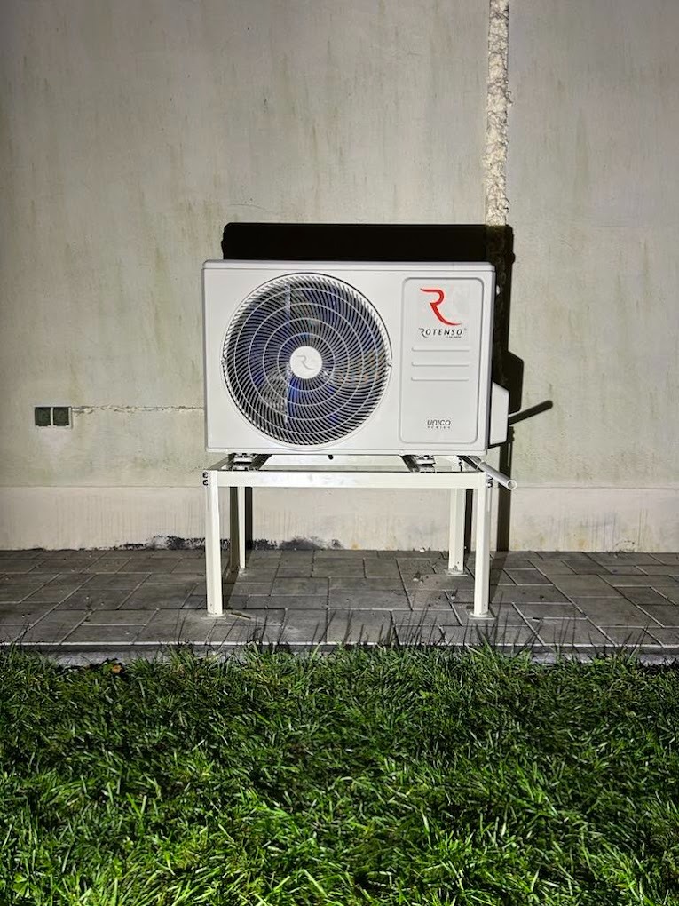 Biała jednostka zewnętrzna klimatyzatora Rotenso Unico Series, umieszczona na metalowym stelażu, stoi na brukowanym podłożu przed ścianą z widocznymi śladami wilgoci i instalacji.