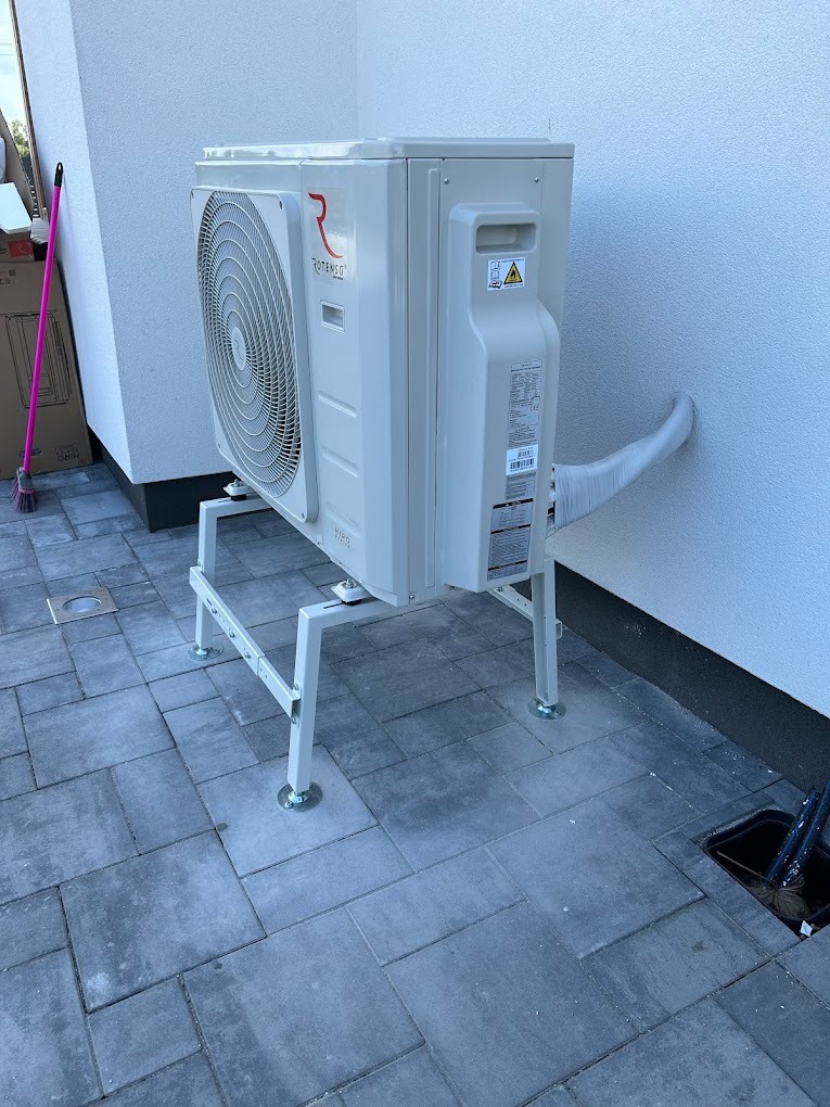 Biała pompa ciepła marki Rotenso zamontowana na metalowym stelażu na szarym brukowanym tarasie, z widoczną rurą prowadzącą do ściany budynku.