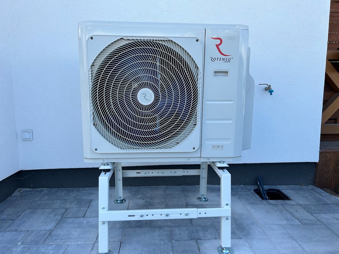 Biała pompa ciepła Rotenso Hiro Series na metalowym stelażu, zamontowana na zewnątrz budynku z jasną elewacją i szarą kostką brukową.