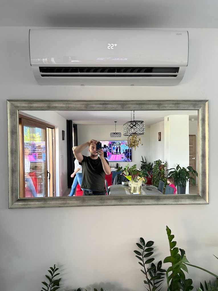 Biały klimatyzator ścienny zamontowany nad lustrem w srebrnej ramie, w którym odbija się osoba robiąca zdjęcie. W tle widoczny salon z roślinami i telewizorem.
