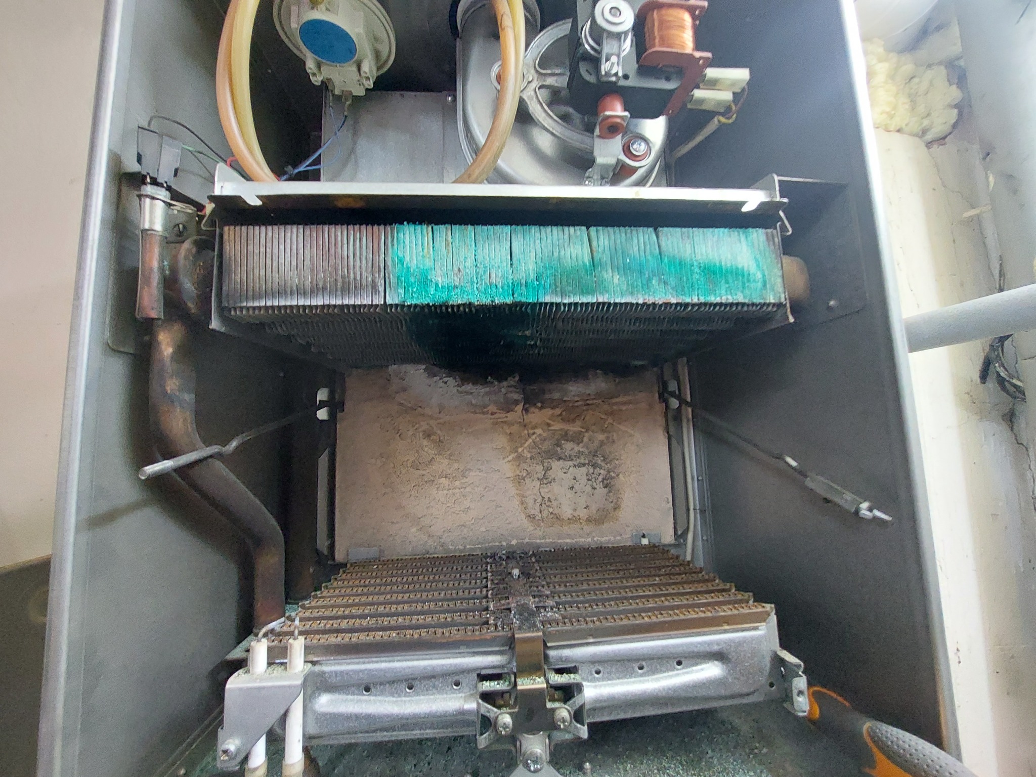 Wnętrze otwartego kotła gazowego z widocznym wymiennikiem ciepła pokrytym zielonkawym nalotem i śladami sadzy, elementy instalacji gazowej oraz fragment ściany z pianką montażową.