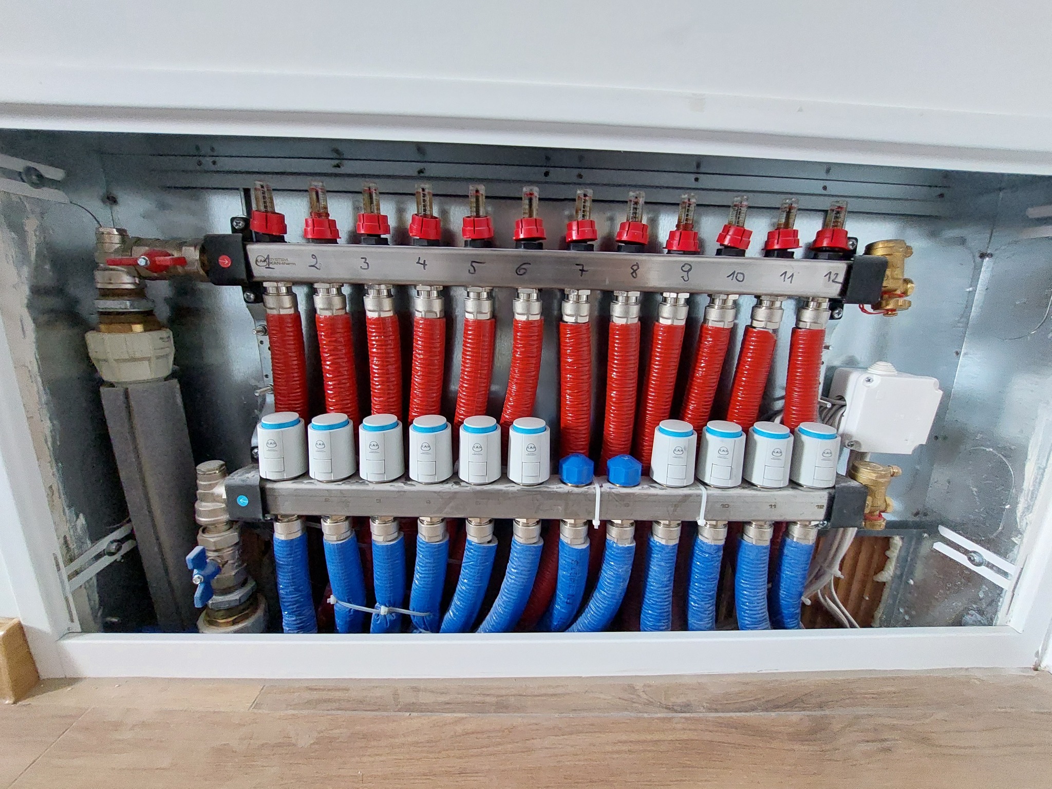 Rozdzielacz ogrzewania podłogowego z czerwonymi i niebieskimi rurami, zamontowany w szafce, widoczny na tle drewnianej podłogi w Sieradzu.
